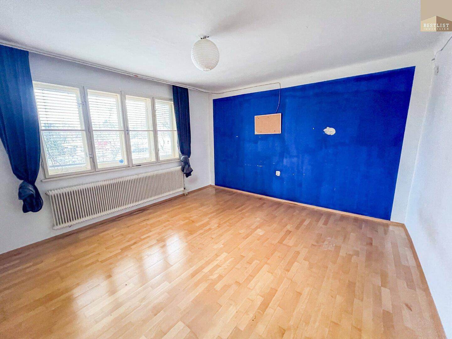 Wohnzimmer mit Holzboden, blauer Akzentwand und großem Fenster mit Heizkörper.