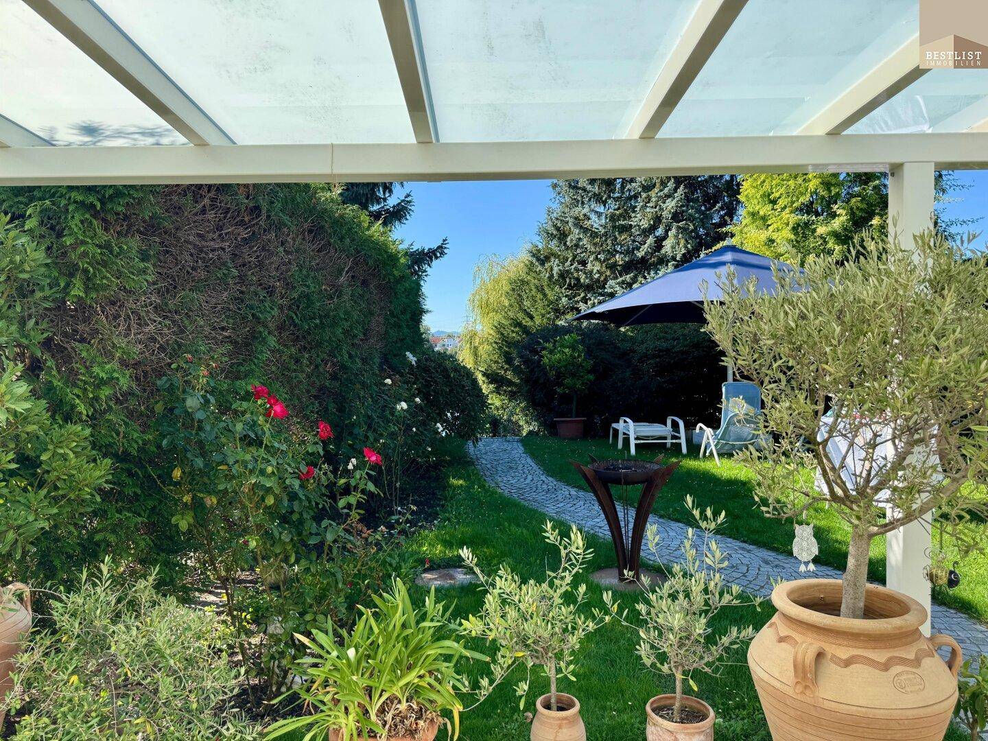 Blick von der überdachten Terrasse in den gepflegten Garten mit blühenden Rosen und einem Feuerkorb.
