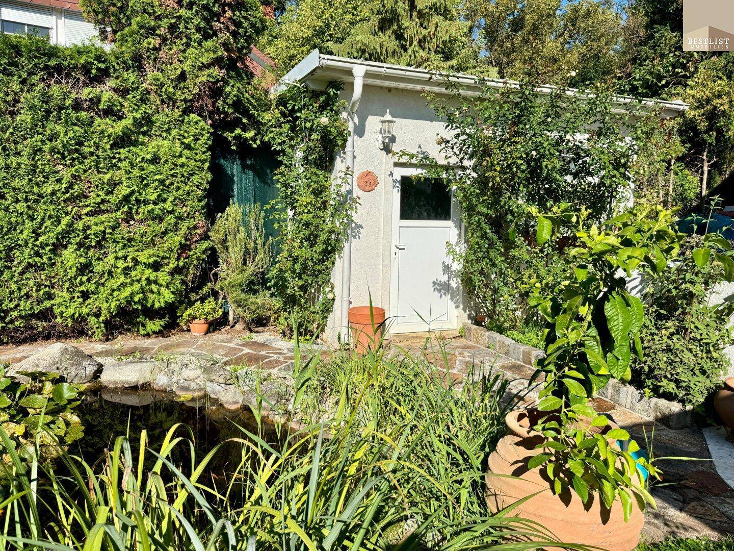 Kleines Gartenhaus umgeben von üppigem Grün und einem Teich, ideal für Gartengeräte.