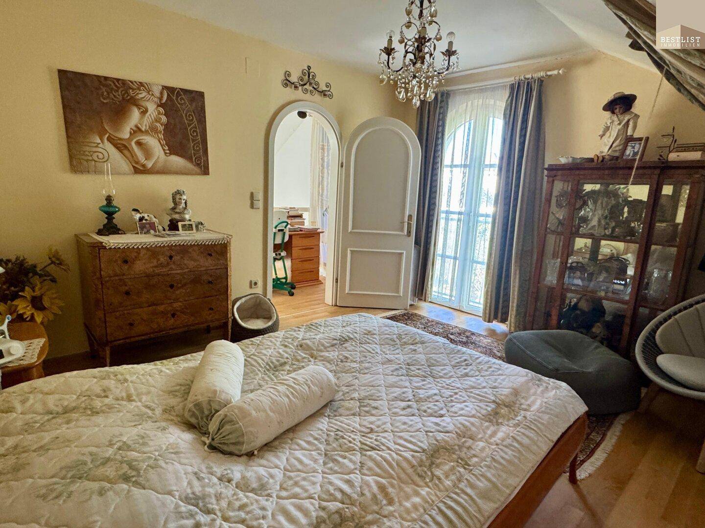 Helles Schlafzimmer mit Doppelbett, Kronleuchter und Zugang zu einem französischen Balkon.