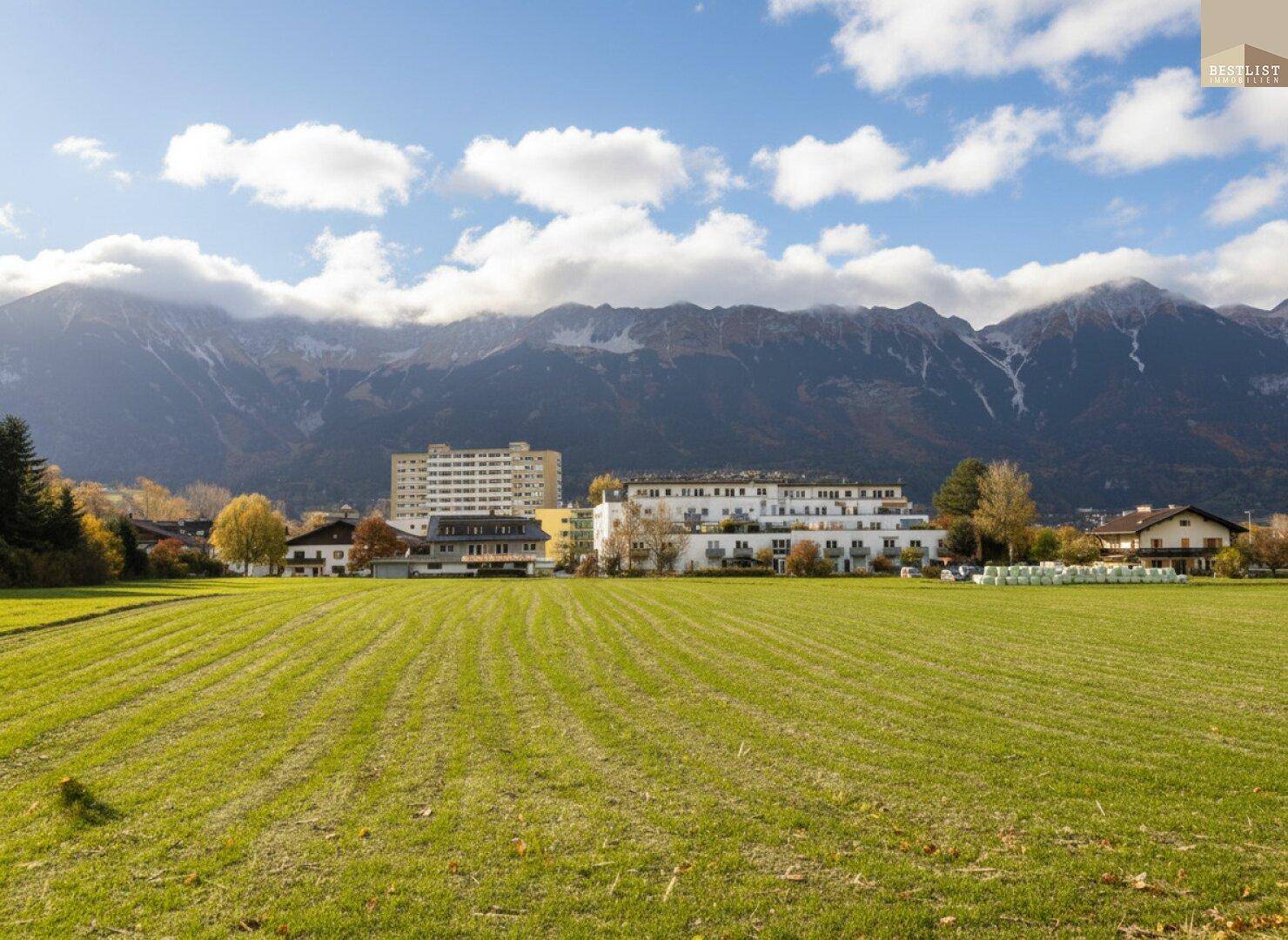 Grüne Wiese mit Wohngebäuden und imposanter Bergkulisse unter strahlend blauem Himmel.