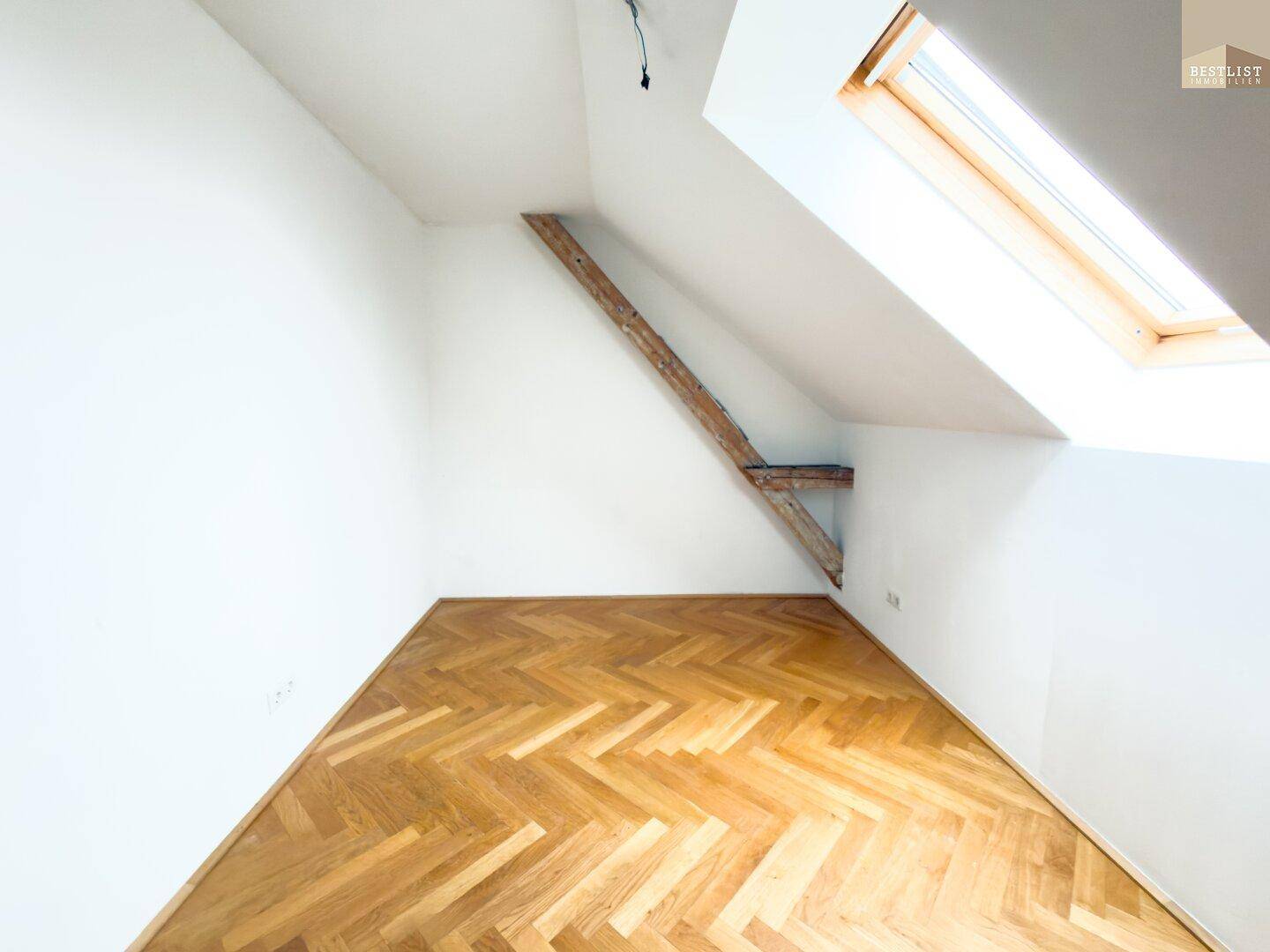 Helles Zimmer im Dachgeschoss mit Dachfenster, Holzbalken und Fischgrätparkett.