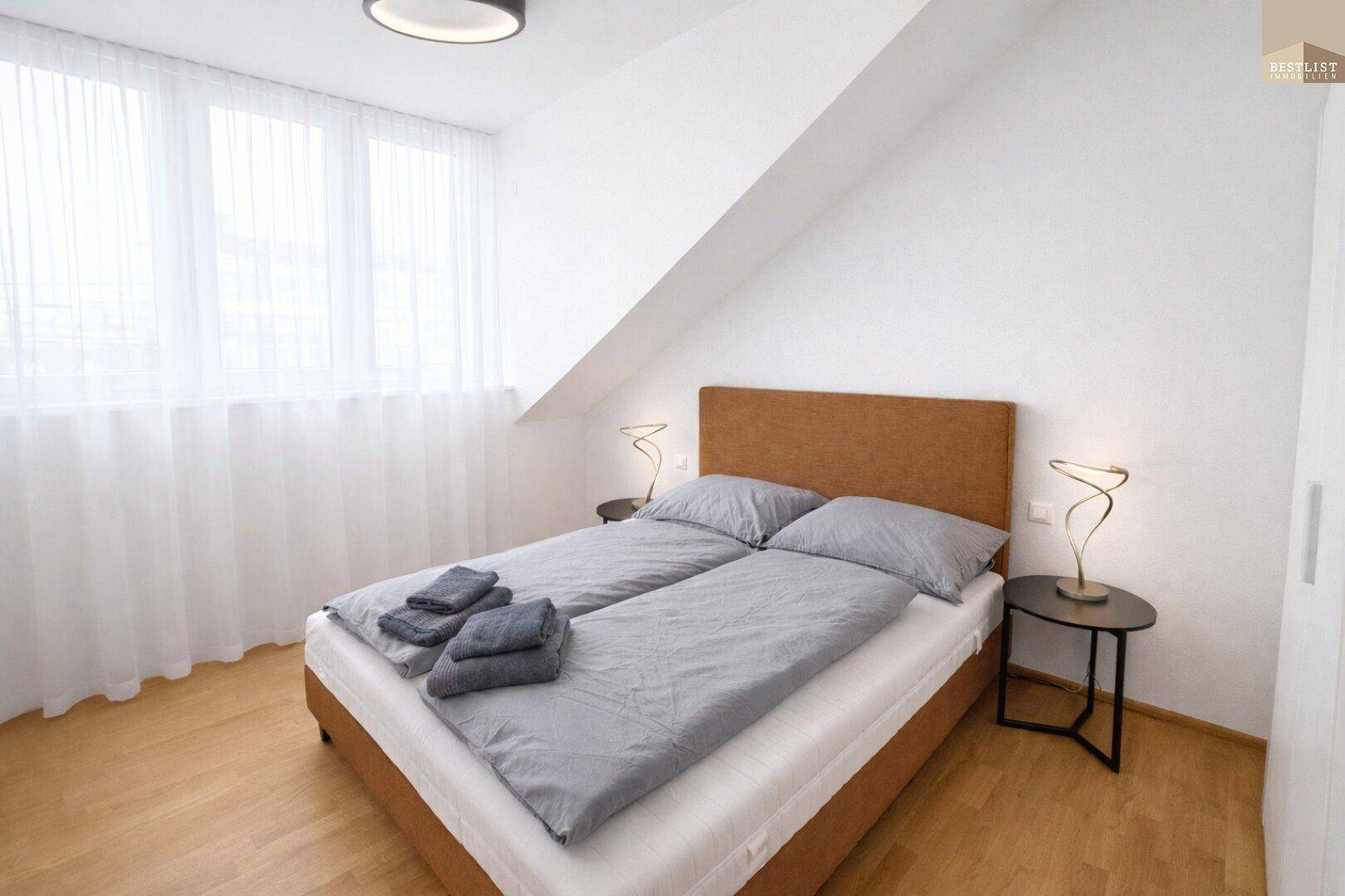 Helles Schlafzimmer mit großem Fenster, Parkettboden und schräger Decke.
