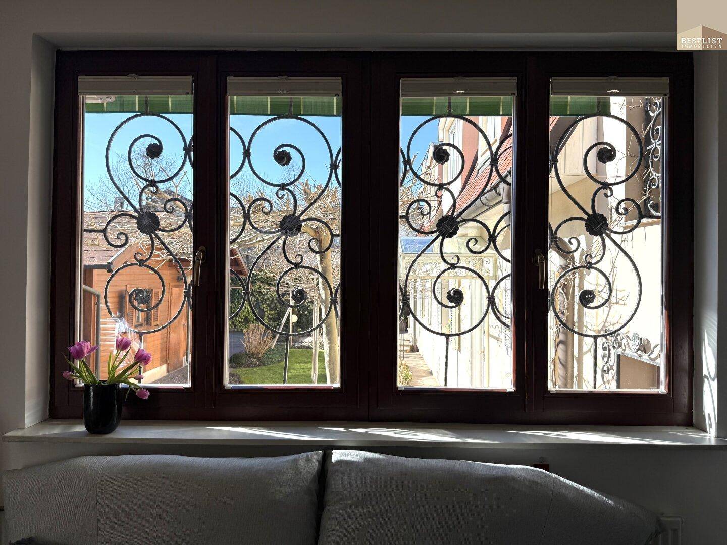 Fenster mit kunstvollen schmiedeeisernen Gittern und Blick in den grünen Gartenbereich.