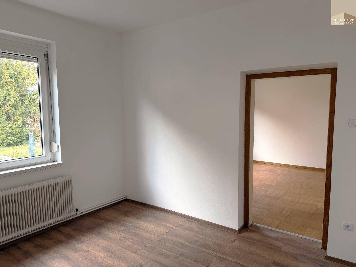 Helles Zimmer mit Laminatboden, weiß gestrichenen Wänden und Fenster mit Blick ins Grüne.