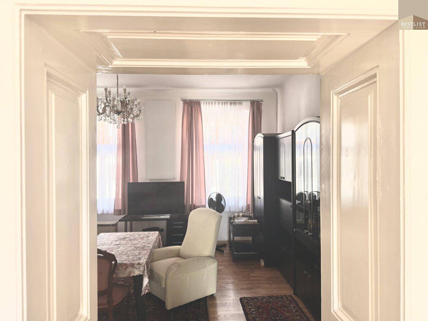 Wohnzimmer mit Kronleuchter, Esstisch und einem Sessel, durch eine Türöffnung sichtbar.
