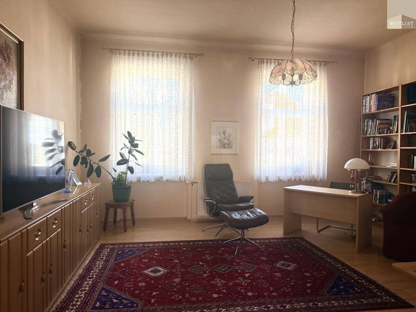 Geräumiges Wohnzimmer mit Teppich, Schreibtisch und Bücherregal, hell durch zwei Fenster.