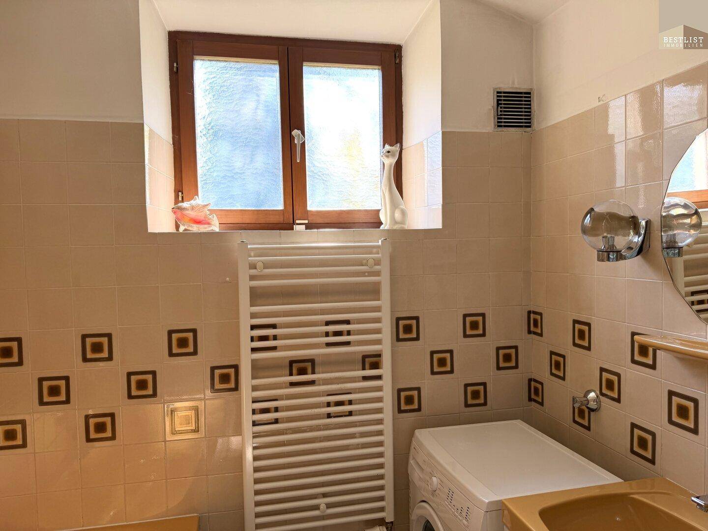 Badezimmer mit beigen Fliesen, Heizkörper und einem Fenster, das Tageslicht hereinlässt.