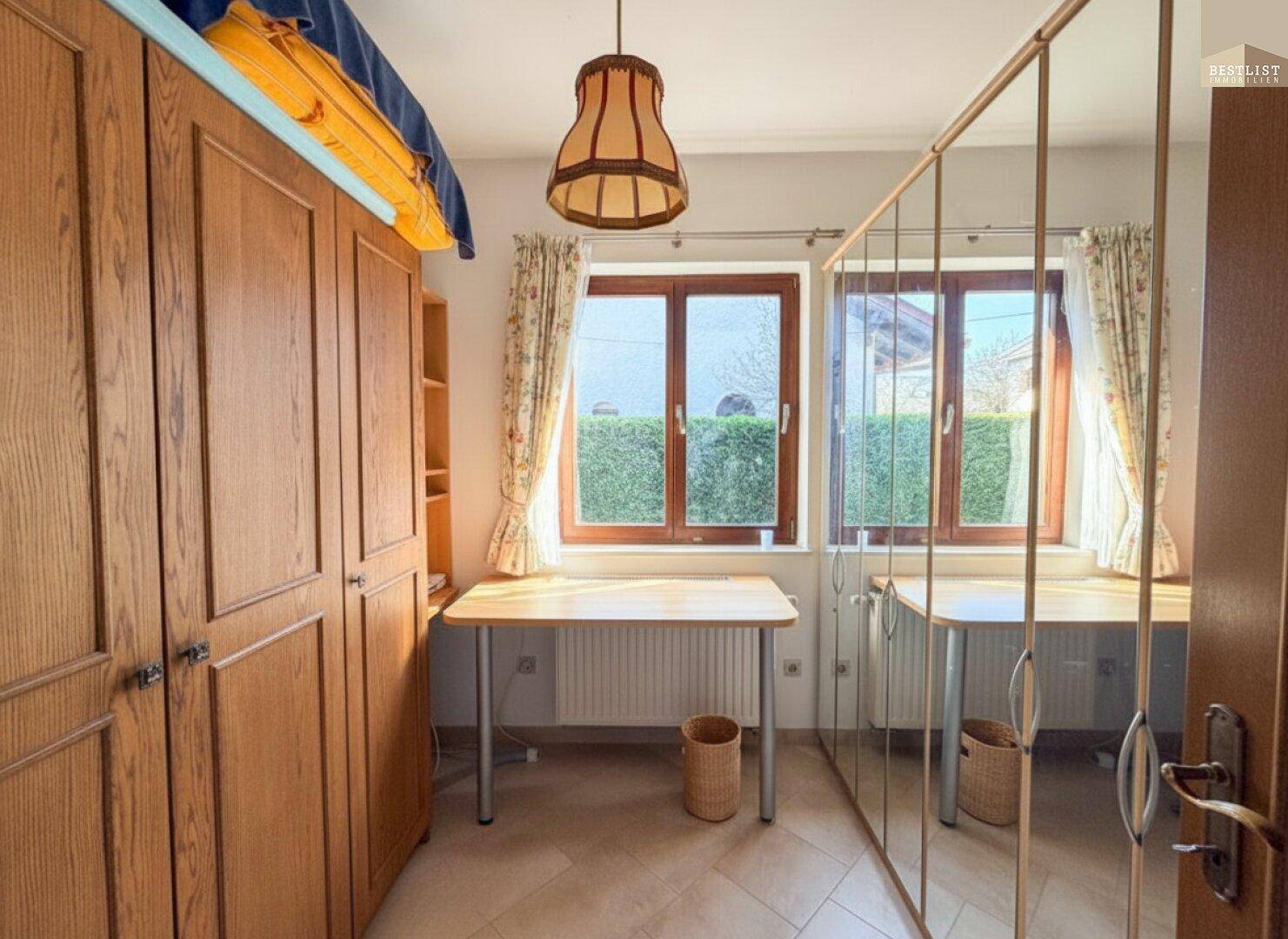 Schlafzimmer mit Holzschrank, Spiegelfronten und Fenster mit Blick auf die Hecke.