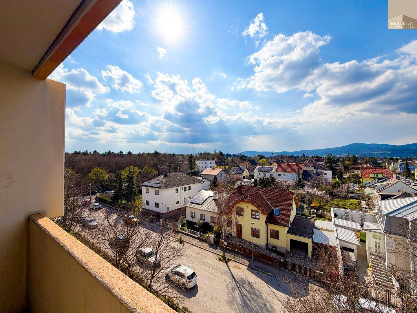 Sonniger Ausblick vom Balkon über eine Wohngegend mit Häusern und Bergen am Horizont.