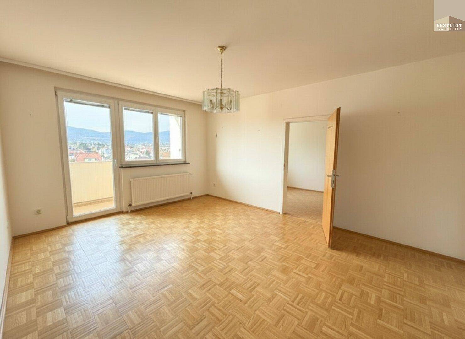 Helles, leeres Wohnzimmer mit Fischgrätparkett, großem Fenster und Zugang zu einem Balkon.