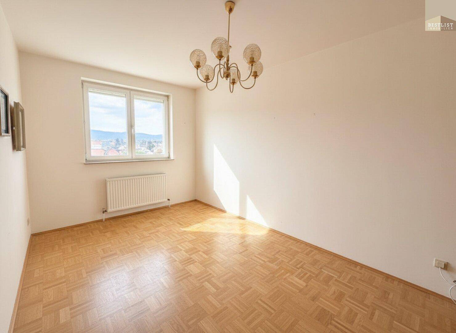 Sonniges, leeres Zimmer mit Parkettboden, Fenster und klassischer Deckenleuchte.
