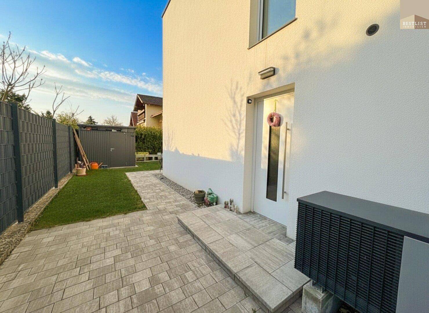 Eingangsbereich des modernen Hauses mit gepflastertem Weg und Wärmepumpe an der Fassade.
