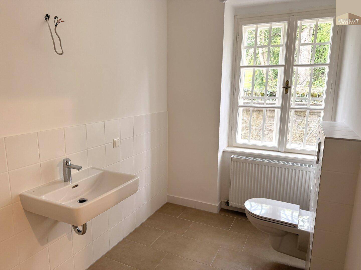 Helles Badezimmer mit Fenster, wandhängender Toilette und Waschbecken, teilweise gefliest.