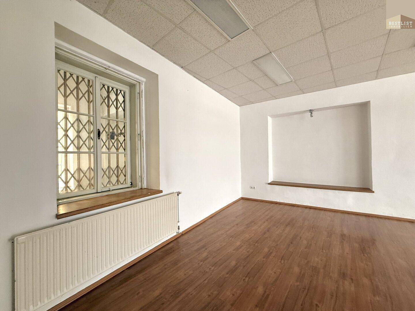 Heller Büroraum mit Holzfußboden, Heizkörper und einem Fenster mit Sicherheitsgitter.