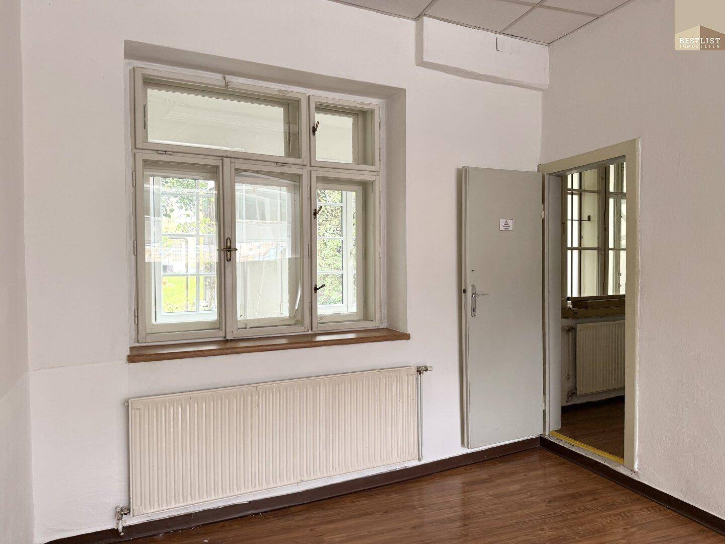Heller Büroraum mit Holzfußboden, Heizkörper und einem Fenster mit Blick ins Grüne.