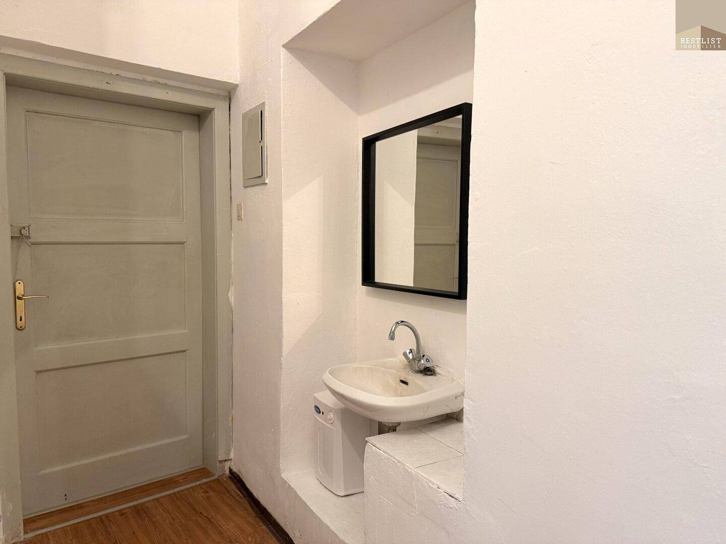 Kleines Badezimmer mit Waschbecken, Spiegel und einer schlichten Holztür.