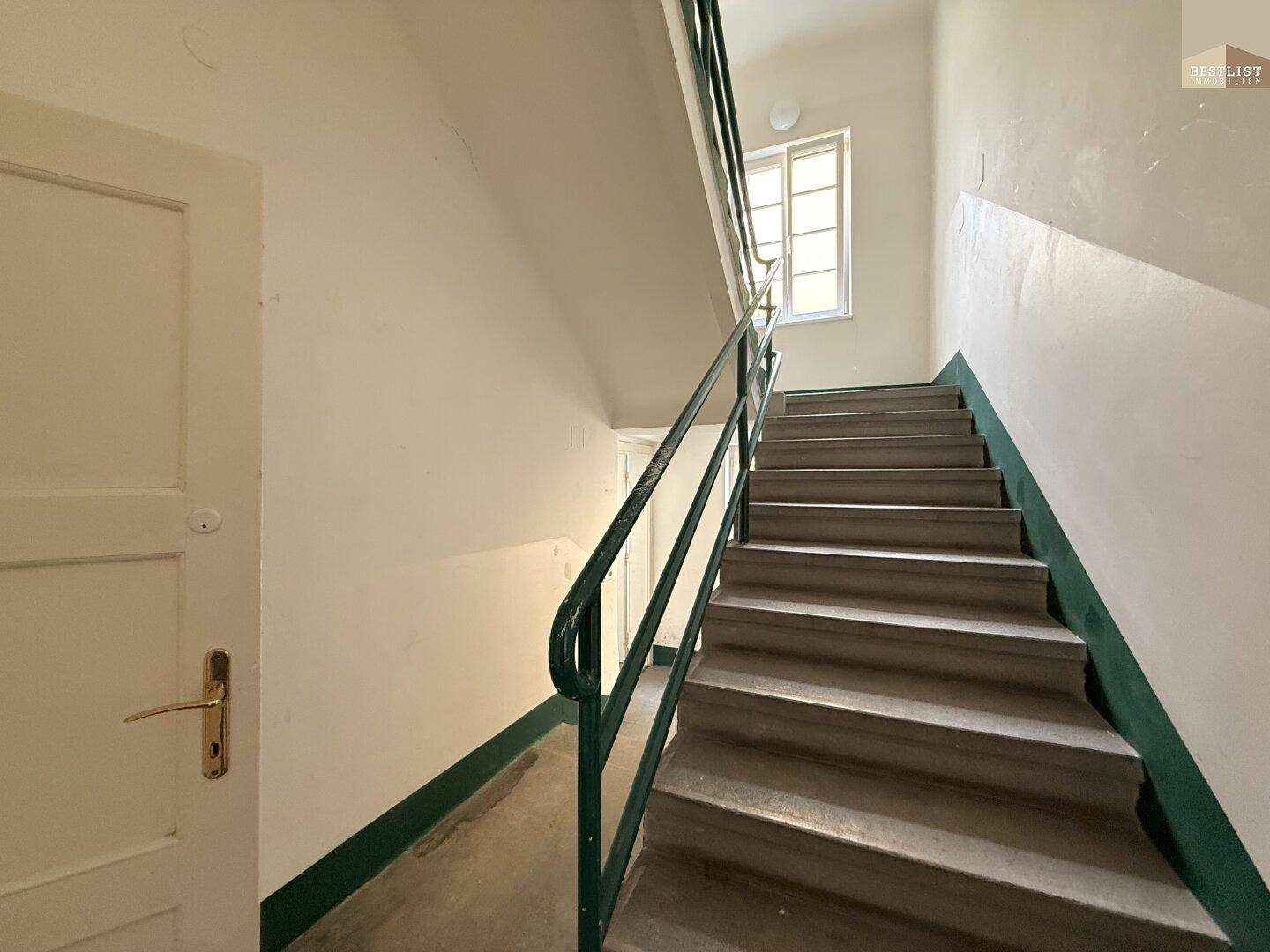 Treppenhaus mit grünem Geländer und einem Fenster, das Tageslicht hereinlässt.