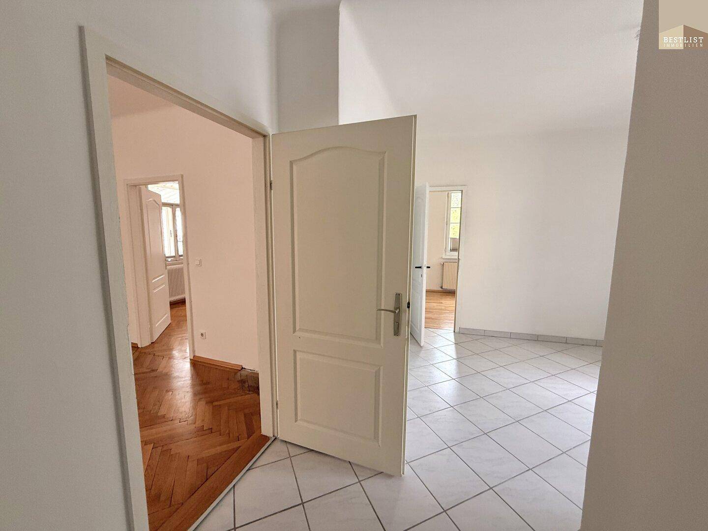 Heller Raum mit Fliesenboden, der zu einem angrenzenden Zimmer mit Parkett führt.
