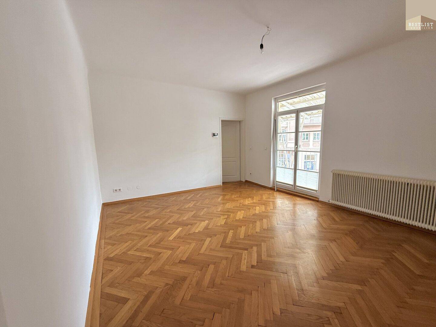 Geräumiges Wohnzimmer mit Fischgrätparkett, Heizkörper und großem Fenster.