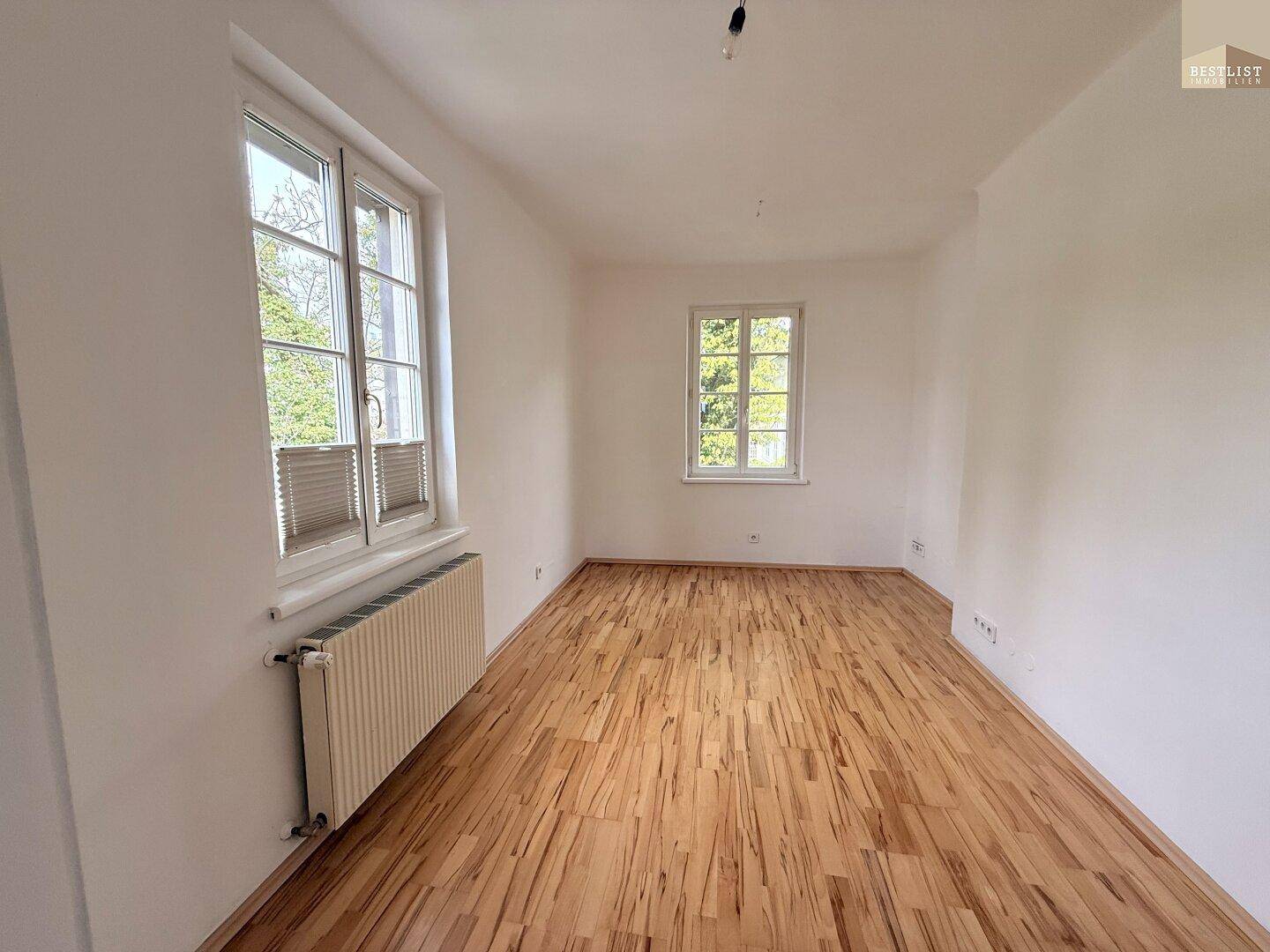 Helles Schlafzimmer mit Holzplankenboden, Heizkörper und zwei Fenstern mit Blick ins Grüne.