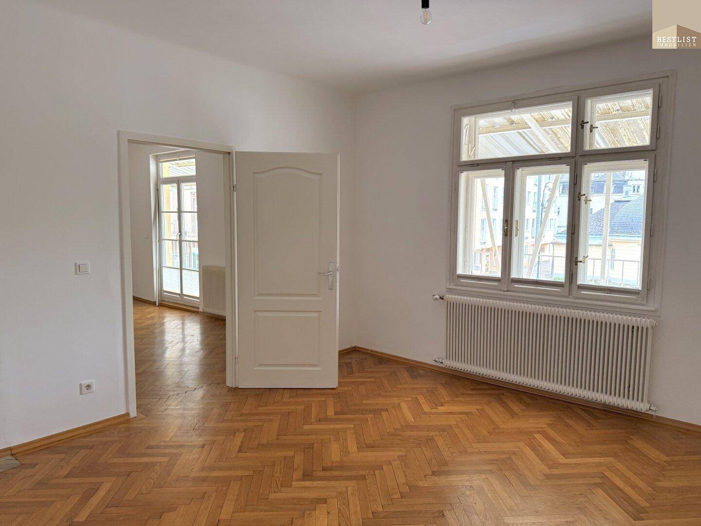 Geräumiges Wohnzimmer mit Fischgrätparkett, Heizkörper und großem Fenster.