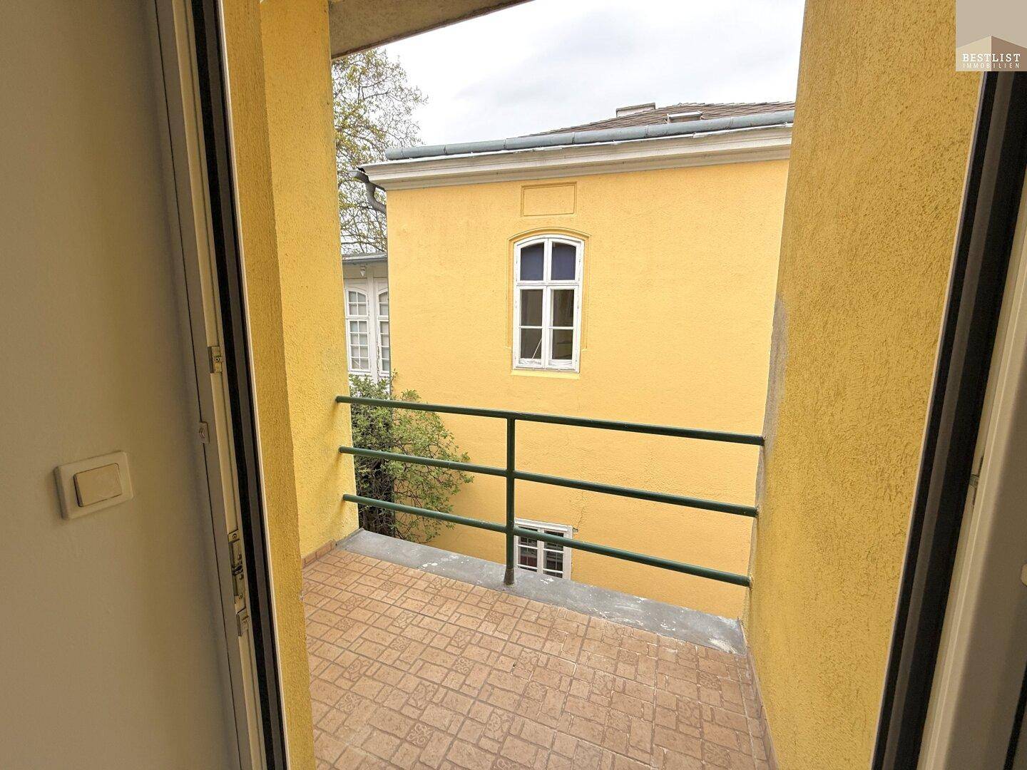 Balkon mit Fliesenboden und grünem Geländer, mit Blick auf ein gelbes Gebäude.