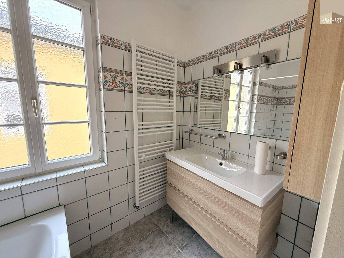 Badezimmer mit Waschbeckenunterschrank, Spiegel, beheiztem Handtuchhalter und Fenster.