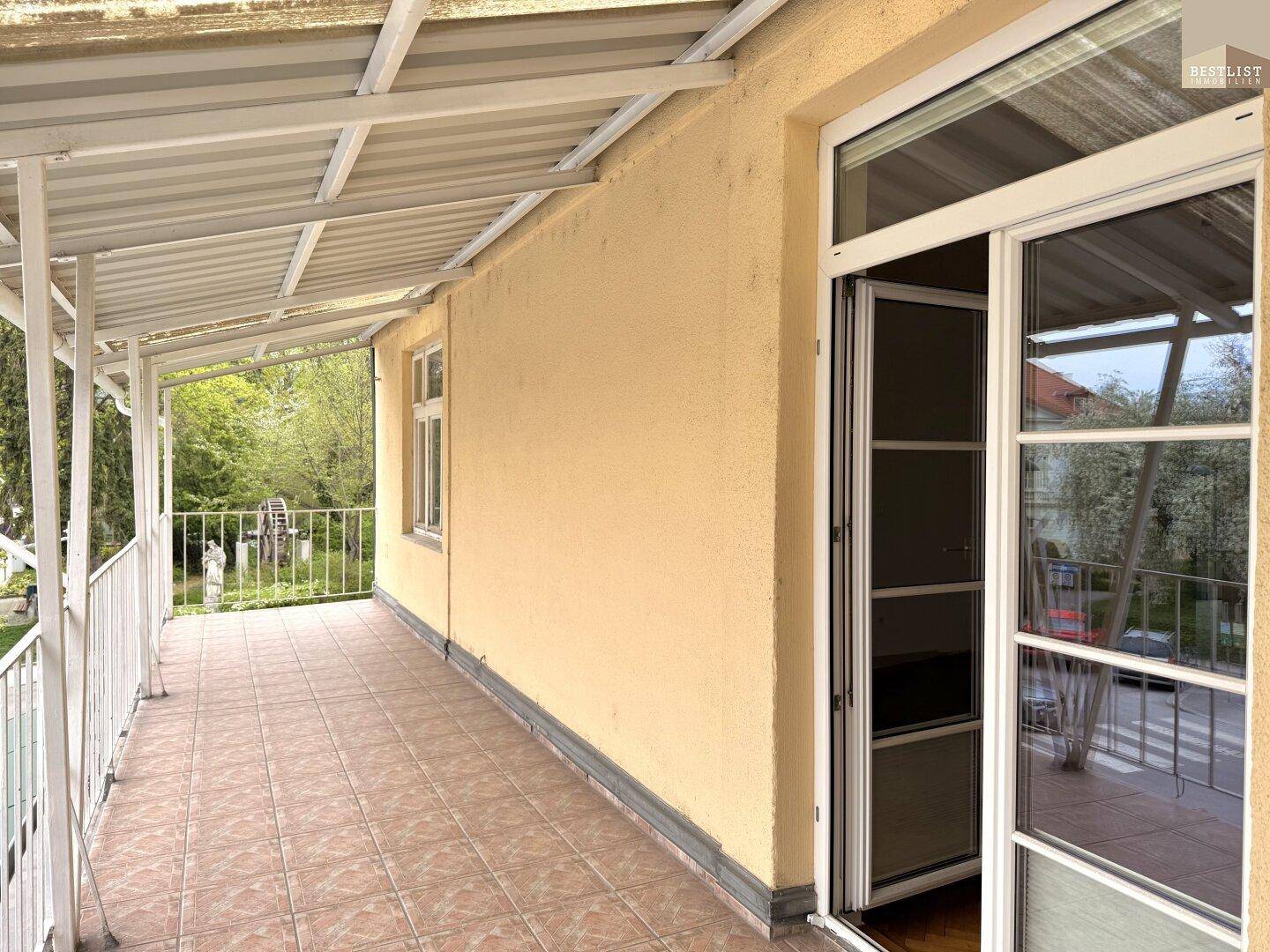 Überdachter Balkon mit Fliesenboden, gelber Fassade und Blick ins Grüne.