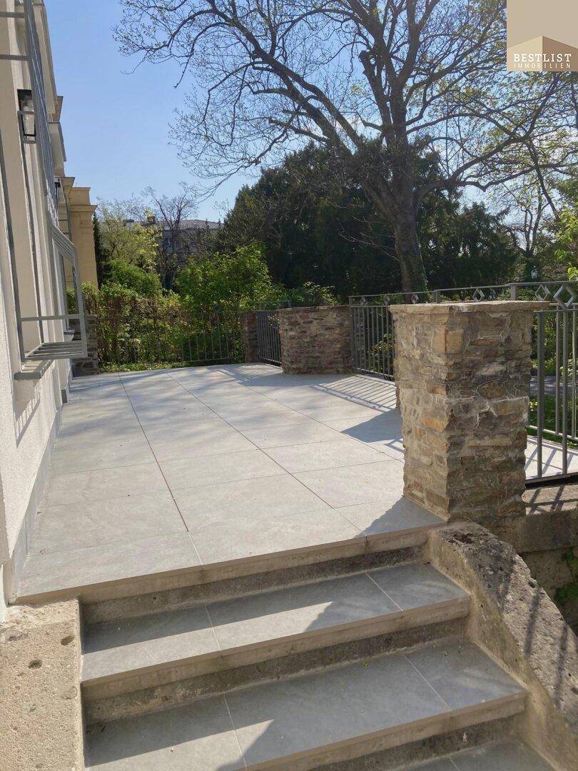 Große geflieste Terrasse mit Steinmauern und Metallgeländer, umgeben von Bäumen.