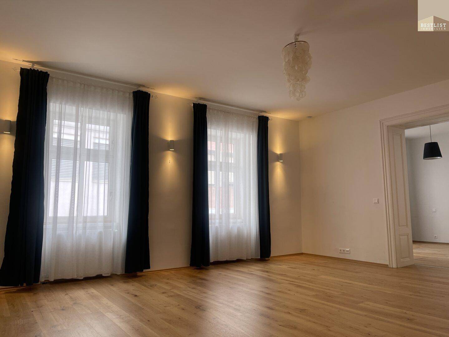 Helles Wohnzimmer mit großen Fenstern, dunklen Vorhängen und Parkettboden.