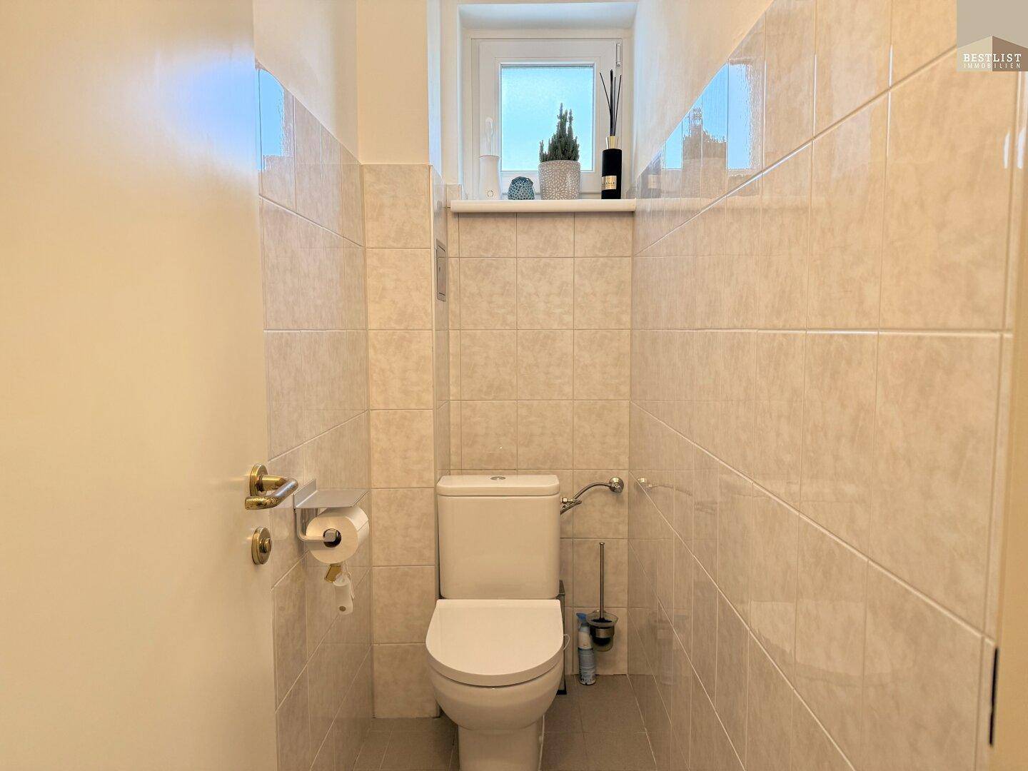 Separates WC mit Fenster, hellen Fliesen und wandmontierter Toilette.