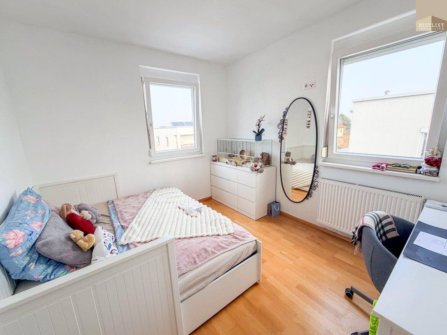 Helles Schlafzimmer mit weißem Bett, Kommode und großem Spiegel, ausgestattet mit Holzboden.