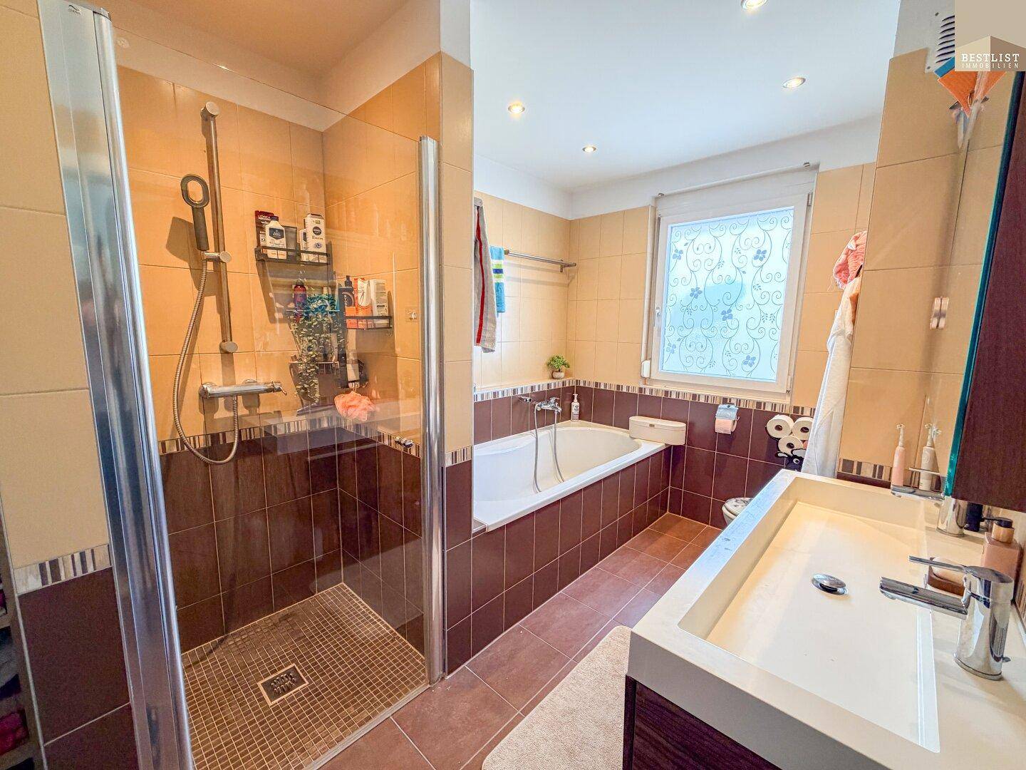 Badezimmer mit braunen und beigen Fliesen, Duschkabine, Badewanne und Fenster.