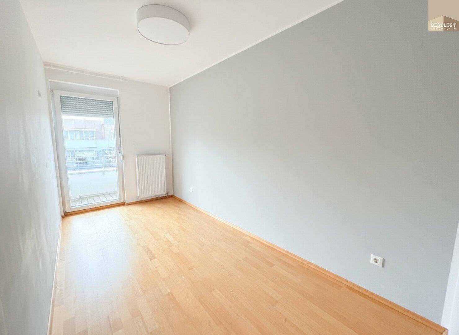 Helles Zimmer mit hellgrauen Wänden, Holzboden und Zugang zu einem Balkon.