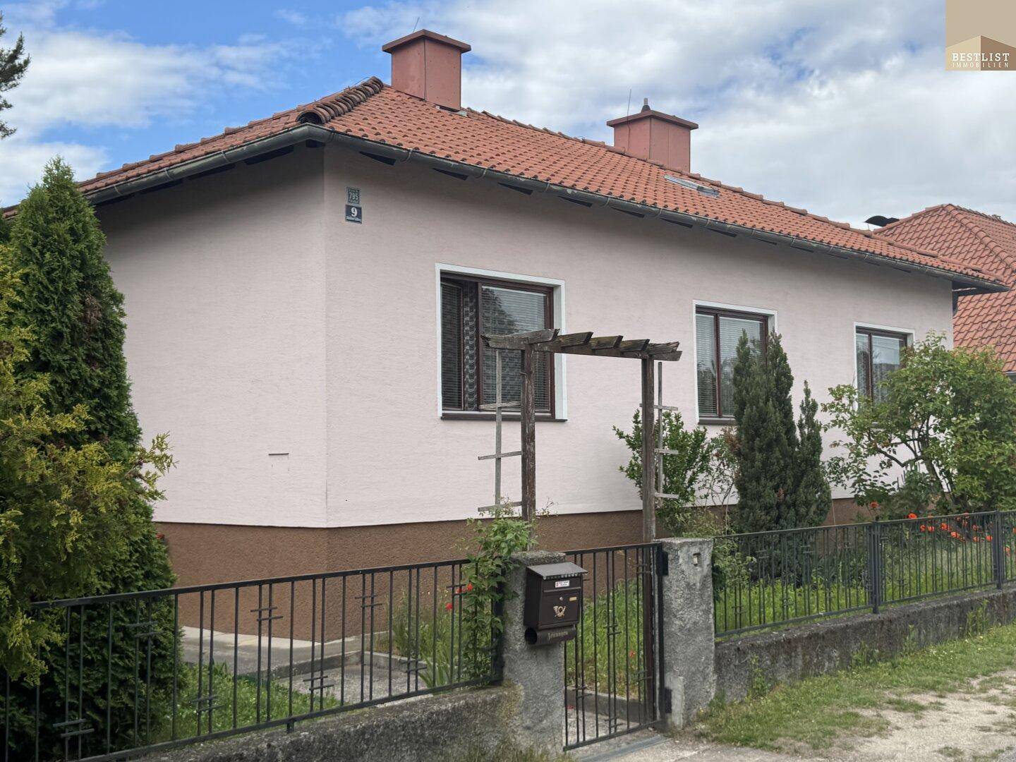 Vorderansicht eines Einfamilienhauses mit rosa Fassade, rotem Ziegeldach und einem kleinen Vorgarten.