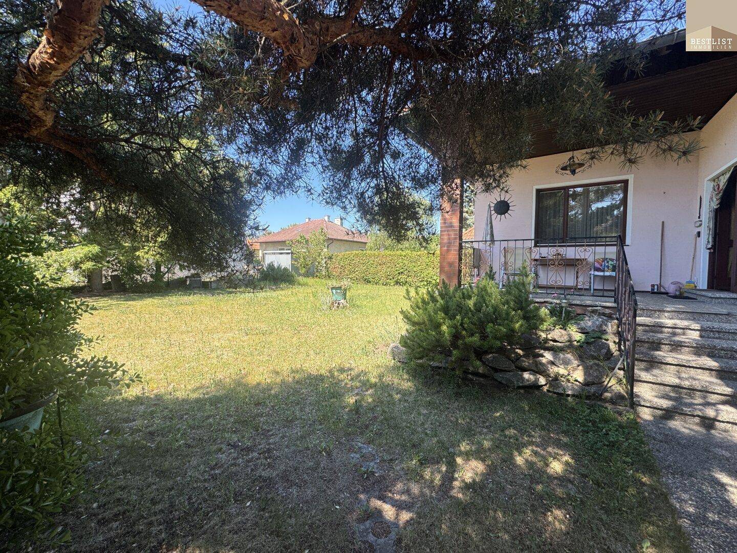 Sonnige Gartenansicht mit altem Baum im Vordergrund und der überdachten Terrasse des Hauses im Hintergrund.