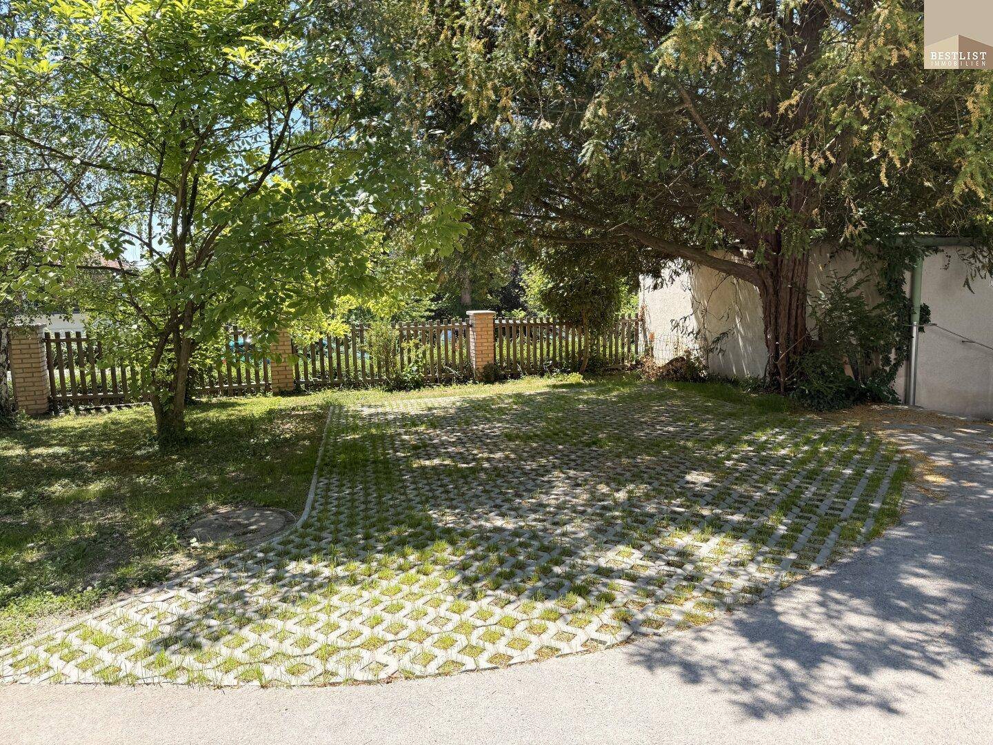 Grüner Parkplatz mit Rasengittersteinen und altem Baumbestand neben einer Garage.