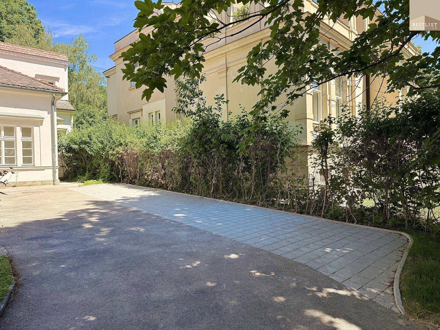 Gepflasterter Parkplatzbereich vor einem hellen Gebäude mit viel Grün und Bäumen.