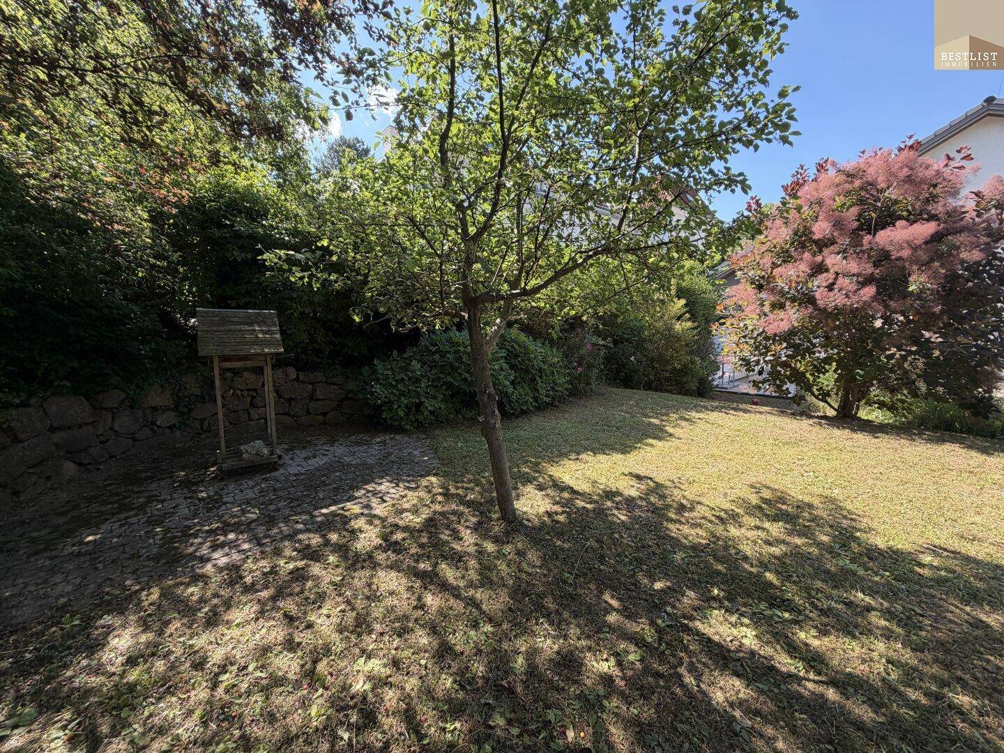 Gepflegter Gartenbereich mit Rasenfläche, Bäumen und einer kleinen Holzhütte unter blauem Himmel.