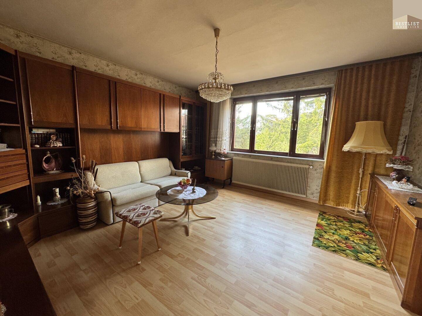Helles Wohnzimmer mit großem Fenster, Laminatboden und traditionellen Holzmöbeln.