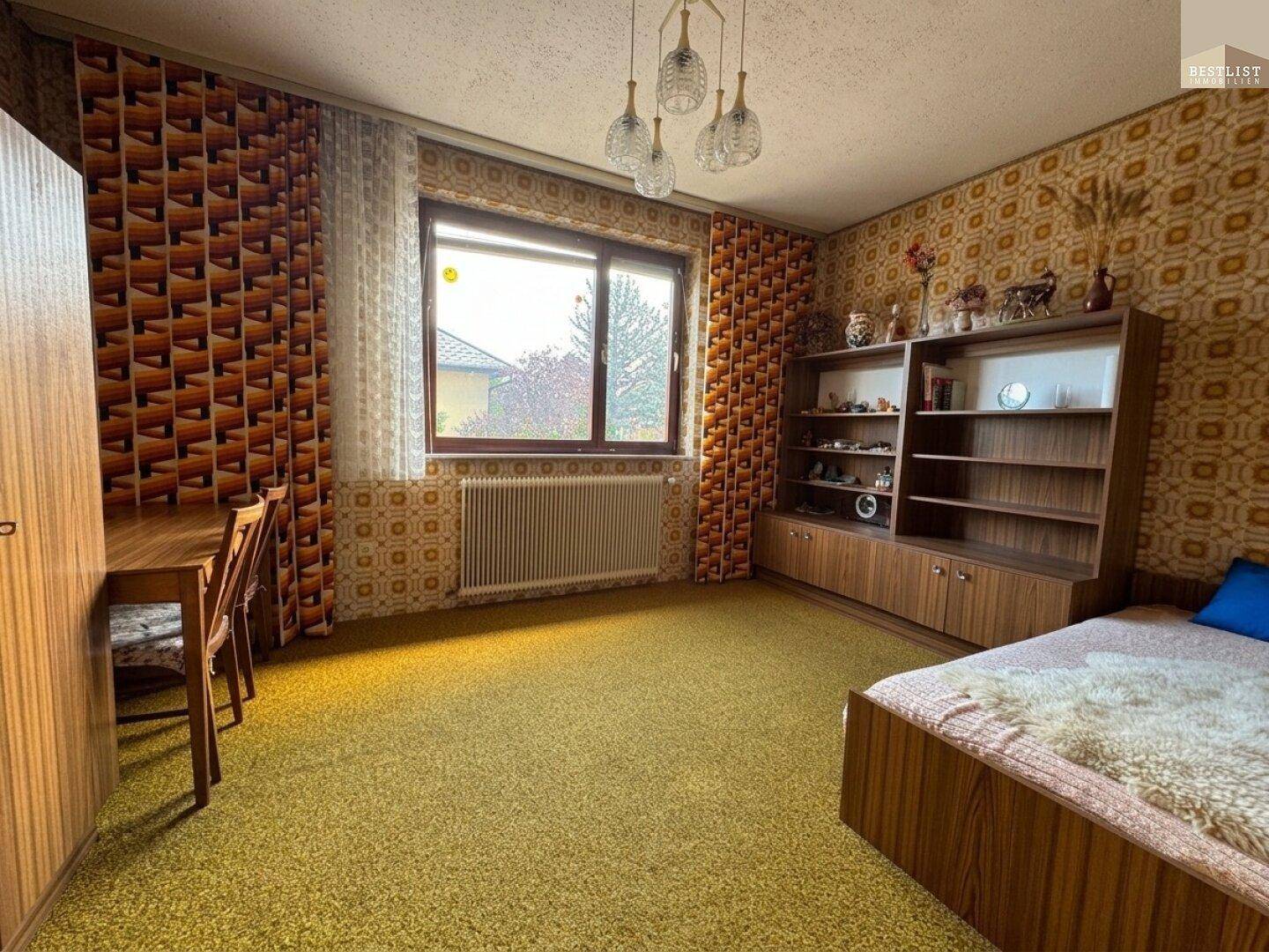Schlafzimmer mit Teppichboden, Holzmöbeln und gemusterter Tapete im Retro-Stil.