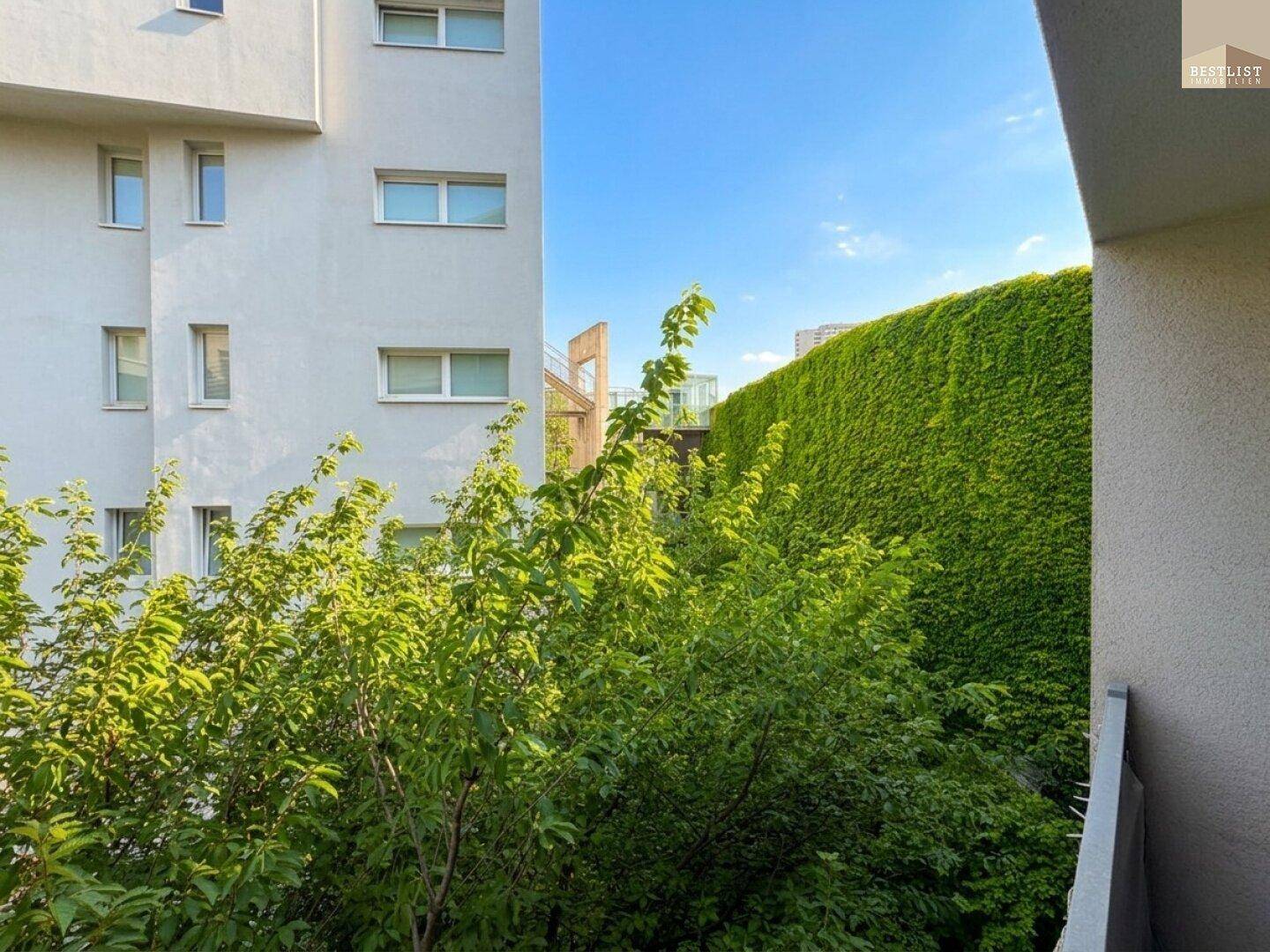 Blick von einem Balkon auf eine grüne Wand und moderne Wohngebäude unter blauem Himmel.