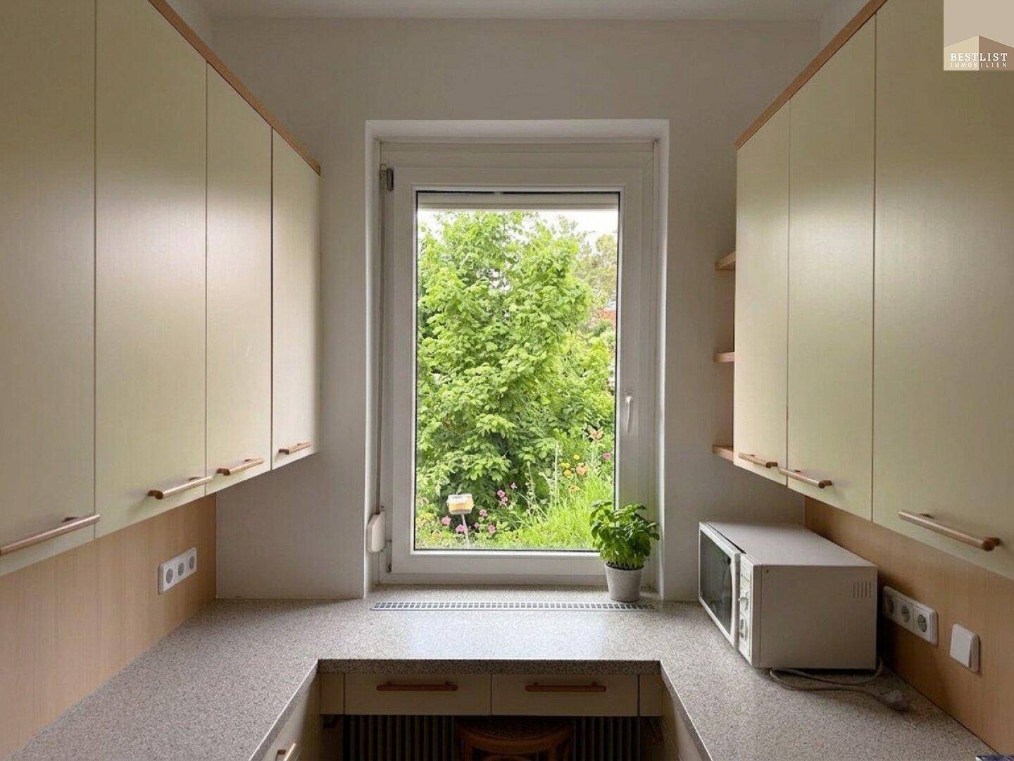 Helle Küche mit beigen Schränken, Arbeitsplatte und Fensterblick ins Grüne.