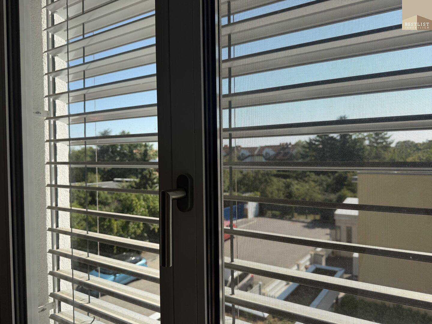 Fenster mit Jalousien bietet einen Blick auf die grüne Umgebung und umliegende Gebäude.