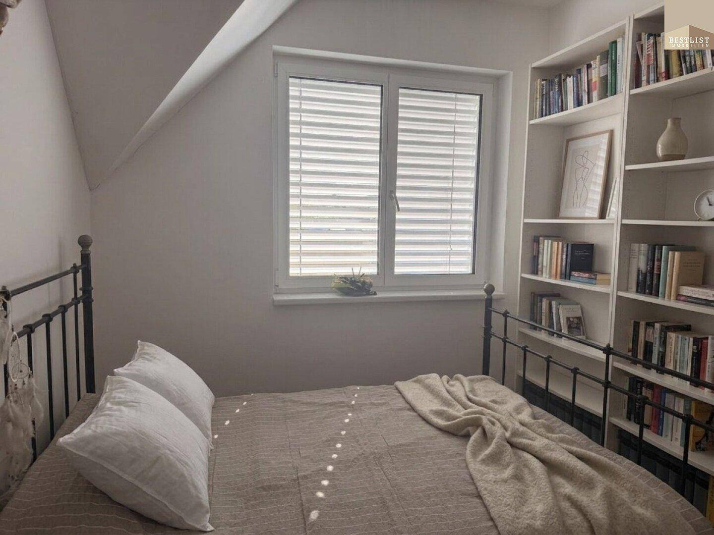 Gemütliches Schlafzimmer mit Metallbett, Bücherregal und Fenster mit Jalousien.