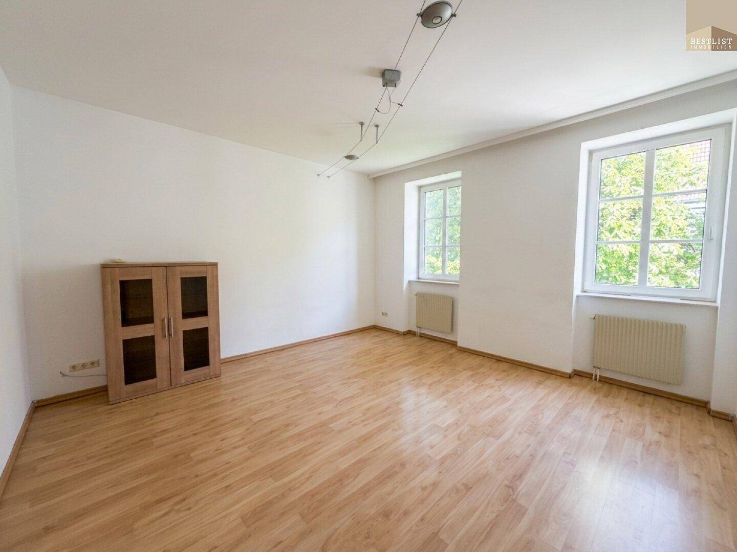 Geräumiges Zimmer mit Holzboden, zwei Fenstern mit Grünblick und einem kleinen Holzschrank.