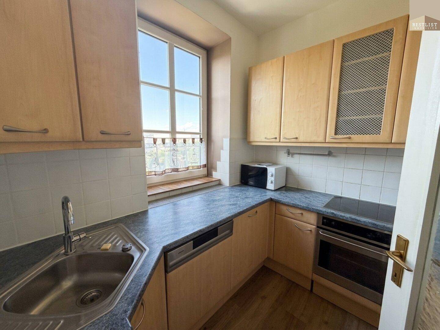 Helle Küche mit Holzschränken, grauer Arbeitsplatte, Spüle, Geschirrspüler und integriertem Backofen.