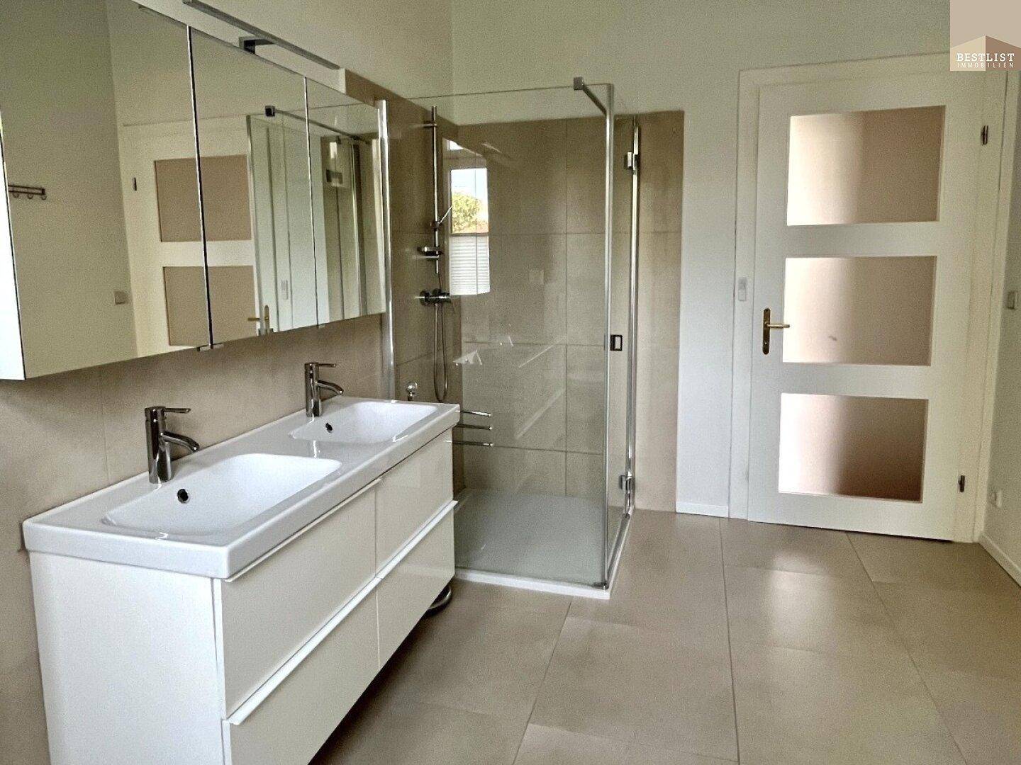 Modernes Badezimmer mit Doppelwaschbecken, großer Glasdusche und hellen Fliesen an Wänden und Boden.