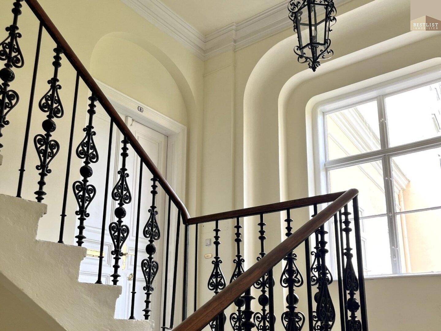 Detailansicht des Treppenhauses mit schmiedeeisernem Geländer, Fenster und Deckenleuchte.